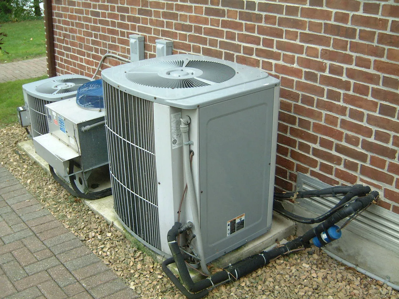 Two central AC condensers on a pad beside a brick house, with insulated refrigerant lines running to the wall